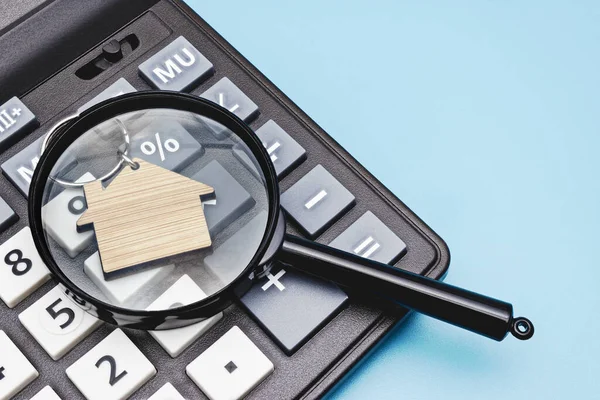 The concept of mortgage, sale and rental of housing and real estate. Mortgage credit lending. Calculator with a wooden house under a magnifying glass on a blue background. Copy space