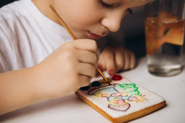Pequeño niño pinta galletas con pinturas de alimentos mientras está sentado en la mesa Hogar concepto de ocio y entretenimiento —  Fotos de Stock