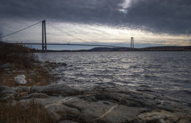 Bulutlu bir gökyüzünün arka planına karşı Rocky sahili ve büyük bir çapa köprüsü