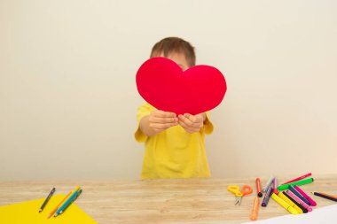 Renkli kağıttan yapılmış kırmızı bir kalp tutan küçük bir çocuk, yüzünü kapatıyor. 8 Mart Anneler Günü konsepti. Kartpostal