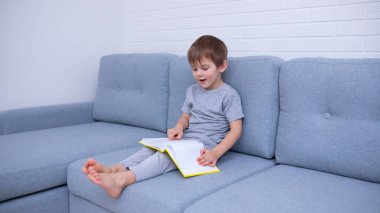 Gri elbiseli küçük bir çocuk gri bir koltukta oturuyor ve sarı bir kitap okumayı öğreniyor. Okuma aşkı, aile zamanı. Kişisel izolasyon, uzaklık öğrenme.
