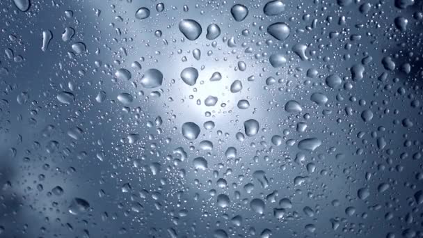 Wet windshield taken from the interior of a car. Rain falls on the surface of a car window glass on a gray background. The sun is shining from behind the clouds. Water drops wallpaper. — Stock Video