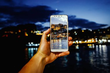 Seyahat ederken akıllı telefonunu cep kamerası olarak kullanıyorsun. Günbatımında turist şehri ekranda