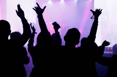 Bir gece kulübündeki konserde el kaldıran insanların siluetleri. Parlak mor ve pembe sahne ışıkları parti atmosferi yaratır..