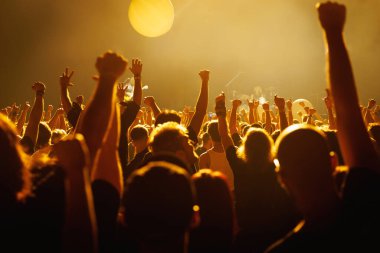 Büyük bir kalabalık konserin keyfini çıkararak, ellerini parlak sahneye kaldırıyor. Altın ışıklandırma ve enerjik atmosfer canlı müziğin ve ortak heyecanın özünü yakalar..