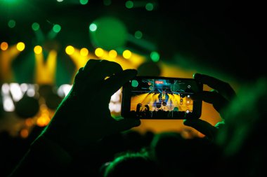 Yeşil ve sarı ışıklar sahneyi aydınlatırken, eller akıllı telefonu bir konseri filme alıyor..