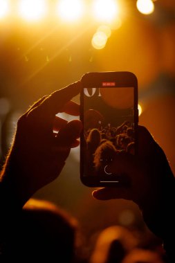 Turuncu-altın ışık altında bir konseri kaydeden akıllı telefonu tutan ellerin yakın çekimi. Çekim, modern eğlence deneyimlerinde bağlantı, heyecan ve teknolojiyi aktarıyor..