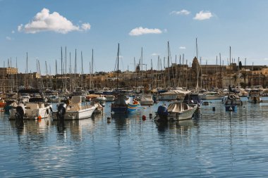 Güneşli bir marinaya demirlemiş yat ve tekne manzarası. Arka planda sakin su ve şehir manzarası var. Seyahat, turizm veya Akdeniz yaşam tarzı kavramları için ideal.
