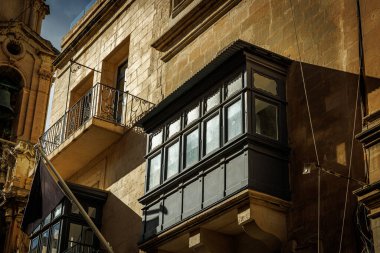 Karanlık ahşap kaplı balkon Valletta 'daki kireçtaşı cephesine gölge düşürüyor. Akdeniz mimarisi ve yerel kültür temalarını resmetmek için harika.
