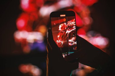 Arka planda kırmızı sahne ışıkları olan akıllı telefon kayıt konser performansını tutan ellerin yakın çekimi.