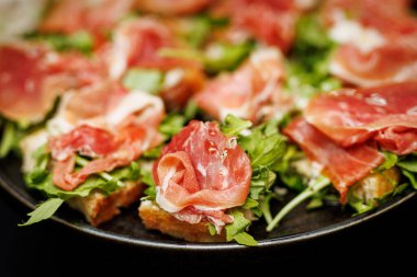 Prosciutto ve roka yapraklarıyla kaplı kızarmış ekmek dilimleri, siyah bir tabakta servis edilir, yakın plan çekimleri yapılır..