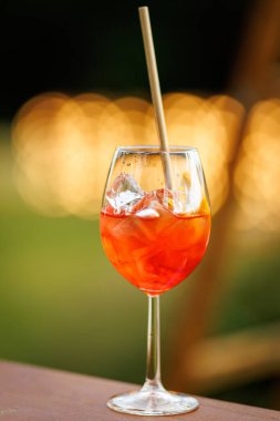 Buzlu turuncu bir kokteylin yakın çekimi ve şarap kadehinde bir pipet, arka planda sıcak bokeh ışıkları ile dışarıda çekilmiş..