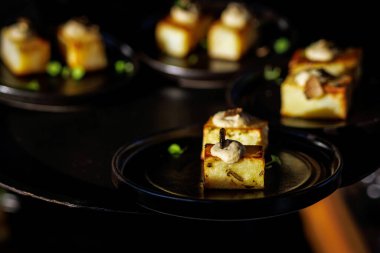 Yer mantarı kremi ve otlarla kaplı küçük kare aperatif parçaları karamsar ışıklandırmada siyah tabaklarda servis edilir..