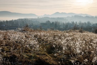 Tepenin yamacındaki buzlu bir tarla arkasında sisli dağlar olan sessiz bir kış köyüne bakıyor. İnce güneş ışığı soğuk sahneye sıcaklık katar..