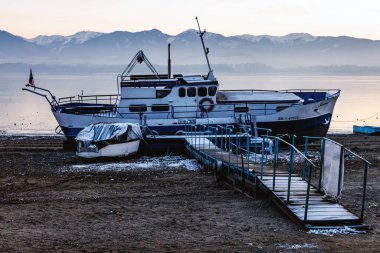 Birkaç tekne, uzak mesafede karla kaplı dağlarla birlikte tahta bir rıhtımın yakınındaki buzlu bir göl kıyısında dinleniyor. Soğuk tonlarda sakin bir kış limanı vurgulanıyor.