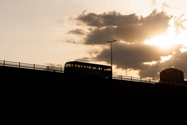 Siluetli otobüs, dramatik günbatımı gökyüzü ve arka planda bulutlarla köprüden geçiyor. Sıcak akşam ışığında şehir taşıma sahnesi.