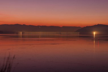 Ilık akşam ışığı, uzak dağları ve parlak ufuk renkleriyle durgun bir göl boyunca yankılanır..