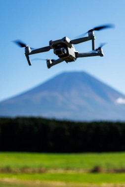 Arka planda Fuji Dağı 'nın bulanık olduğu bir kuadkopter insansız hava aracının yakın çekimi. Sahne, modern hava ekipmanları ve açık hava kullanımını vurguluyor.