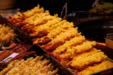 Karides tempurasıyla dolu tepsiler bir Japon sokak yemekhanesinde sergileniyor. Ilık ışıklandırma, kızarmış altın hamuru ve davetkar görünümü arttırır..