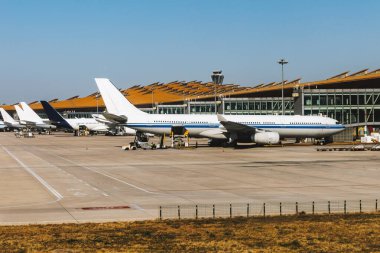 Çok sayıda ticari uçak açık mavi gökyüzünün altında büyük bir havaalanı terminaline park edilmiş, yer ekipleri uçakların etrafında çalışıyor..