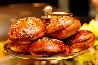 Üzerinde susam ve siyah tohumlar olan parlak mini hamburger çörekleri altın bir stantta sunuluyor. Yemek ve etkinlikler için taze ekmek..