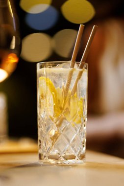 Uzun bir kristal bardakta servis edilen limon dilimleri ile ferahlatıcı, berrak bir içecek. Bokeh ışıkları sıcak bir akşam ortamı ekler..