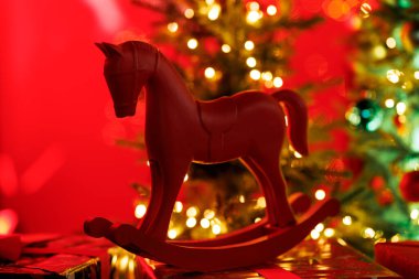Geleneksel, stilize kırmızı sallanan at oyuncağı, parlak ışıklandırılmış bir Noel ağacının bulanık arka planına karşı sarılı bir hediye kutusunun üzerinde duruyor..