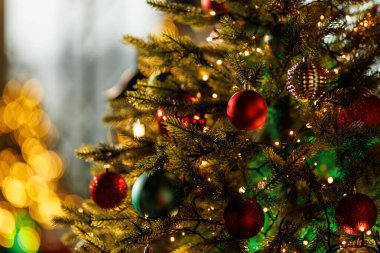 Geleneksel yuvarlak kırmızı ve koyu yeşil süslemelerle süslenmiş gür, yapay bir Noel ağacının yakın plan görüntüsü ve sıcak, parlak ip ışıkları..