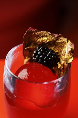 Üzerinde böğürtlen ve metalik altın yaprak olan kırmızı bir kokteylin yakın çekimi. İçki canlı bir renk ve dekoratif bir sunumla gösteriliyor..