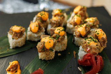 Isırık büyüklüğünde suşi ruloları baharatlı ton balığı karışımı ile kaplanır ve siyah bir sayfanın üzerinde yeşil yapraklar üzerinde sıralanır..