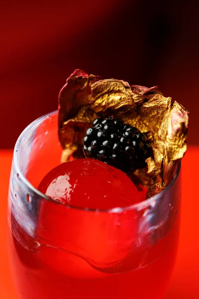 Üzerinde böğürtlen ve metalik altın yaprak olan kırmızı bir kokteylin yakın çekimi. İçki canlı bir renk ve dekoratif bir sunumla gösteriliyor..