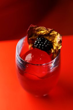 Üzerinde kurumuş çiçek yaprağı ve böğürtlen bulunan büyük bir buz küresi olan canlı kırmızı bir içecek. Parlak arkaplan dramatik renk karşıtlığını artırır.