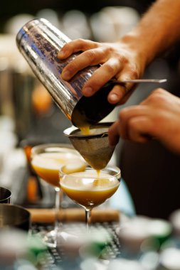Bir barmen ince metal süzgeçten karıştırılmış bir içeceği coupe bardağına doldurur. Barda tam bir kokteyl hazırlama anı..