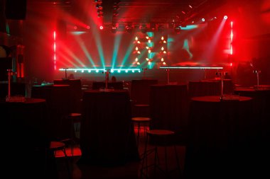 Loş ışıklı bir salon, uzun yuvarlak masalar, tabureler ve güçlü kırmızı ve deniz mavisi sahne ışıklarının yer aldığı bir etkinliğe hazırlanıyor. Atmosfer, yaklaşan bir performans ya da toplanma olduğunu gösteriyor..