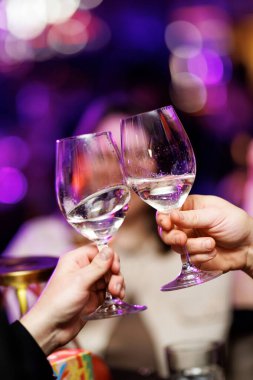 Canlı mor bokeh ışıklarıyla şenlikli bir atmosferde iki kişi şarap kadehlerini kaldırıp tokuşturuyor. Sahne, bir akşam etkinliğinde kutlama anı yakalar..