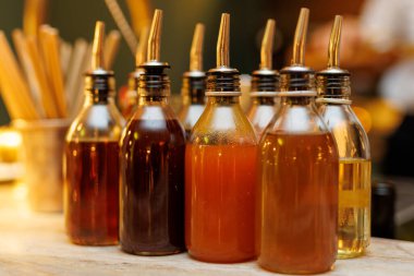 Farklı renklerde yağlarla dolu çoklu cam şişeler ahşap bir masanın üzerine dizilmiştir. Sıcak ortam aydınlatması rahat bir atmosfer yaratır..