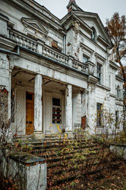 Kötüleşen tarihi bir bina çatlak sütunlar, soyulmuş boyalar ve tahta pencereler gösteriyor. Yapı yıpranmış ve ihmal edilmiş görünüyor, aşırı gelişmiş bitkilerle çevrili..