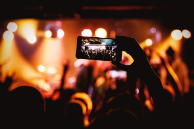 Kalabalık ve sahne ışıkları arka planda bulanıklaşırken, bir akıllı telefon canlı konser kaydı yapıyor..