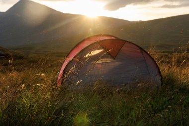Küçük kamp çadırı, tepelerin arkasındaki sıcak günbatımı ışığıyla aydınlatılan bir dağ çayırında uzun otların arasında duruyor..