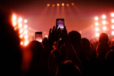 Kalabalık dinleyiciler, canlı performansı yakalamak için kollarını ve telefonlarını kaldırıyorlar. Sahne sıcak turuncu spot ışıkları ve sahne alevleriyle aydınlatılıyor..