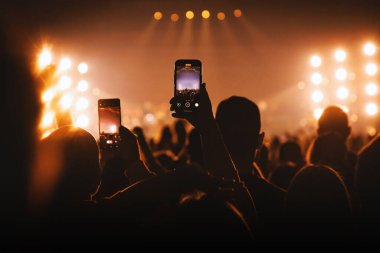 Canlı bir etkinlikte büyük bir seyircinin silueti. Merkezde bir kişi var. Işıklı sahnenin videosunu çekmek için bir telefon tutuyor..