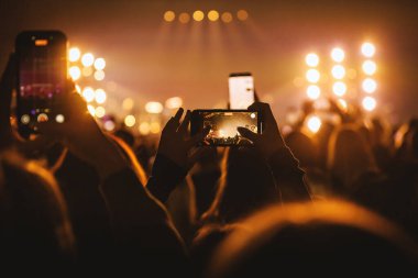 Kalabalığın yakın çekimi, bir festival sırasında parlak bir müzik sahnesini kaydetmek için akıllı telefonu yatay olarak tutan kişiyi gösteriyor..