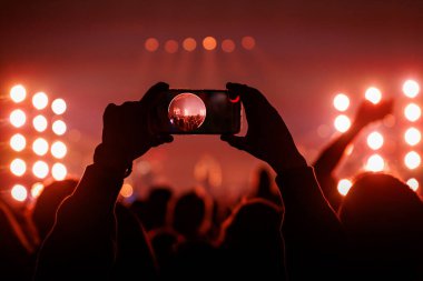İki elle tutulan bir cep telefonunun merkez odağı ekranında sahnenin net bir görüntüsünü gösteriyor. Çevresindeki konser ortamı koyu kırmızı ve sahne ışıkları ile karanlıktır..