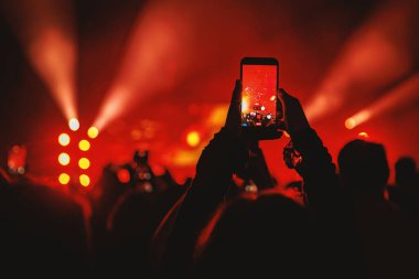 Parlak kırmızı ışık huzmelerinin yer aldığı bir konser sahnesini yakalamak için cep telefonunu dikey olarak tutan bir kişinin yakın plan silueti..