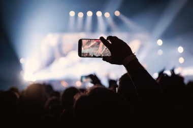 Bir müzisyenin yakın çekimini gösteren cep telefonu ekranına odaklan. Konser salonu serin mavi ışık ve sisli atmosferle dolu..
