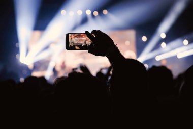 Seyircilerden biri, yoğun mavi ve beyaz sahne ışıkları altında çalan bir grubu kaydetmek için akıllı telefon kullanıyor..