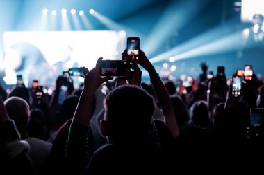Siluetli seyirciler bir müzik festivalinde canlı performans yakalamak için cep telefonlarını kaldırıyorlar. Sahne dramatik mavi sahne ışıkları ve spot ışıklarıyla aydınlatılıyor..