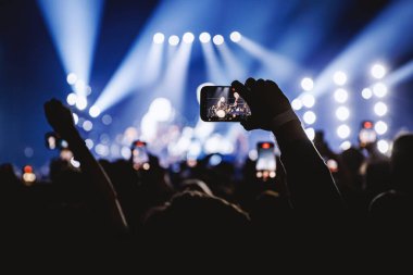 Karanlık bir kalabalığın içindeki bir kişi mavi ve beyaz spot ışıklarıyla aydınlık bir müzik sahnesini kaydetmek için cep telefonunu yatay olarak tutuyor..