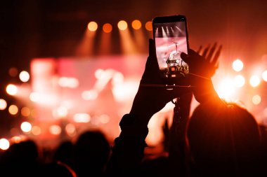 Bir cep telefonu ekranında, parlak turuncu ve kırmızı spot ışıklarıyla aydınlatılmış bir sahnede şarkı söyleyen bir sanatçının kaydı yer alıyor..