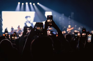 Kalabalıktaki birçok taraftar mavi ve beyaz sahne ışıkları altında canlı yayın yapmak için akıllı telefonları tutuyor..